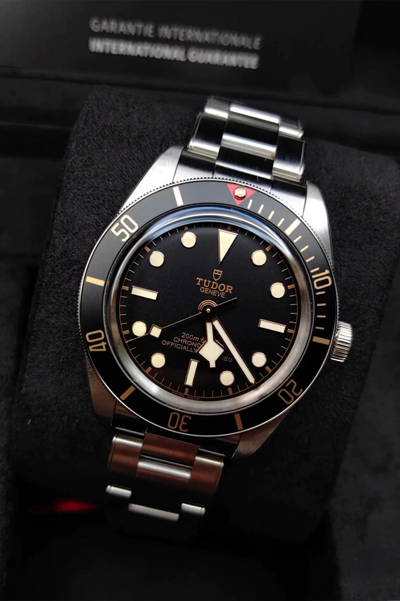 A TUDOR Black Bau watch with a black dial, black rotating bezel, and a silver-toned stainless steel Oyster bracelet, presented inside a black watch box.
