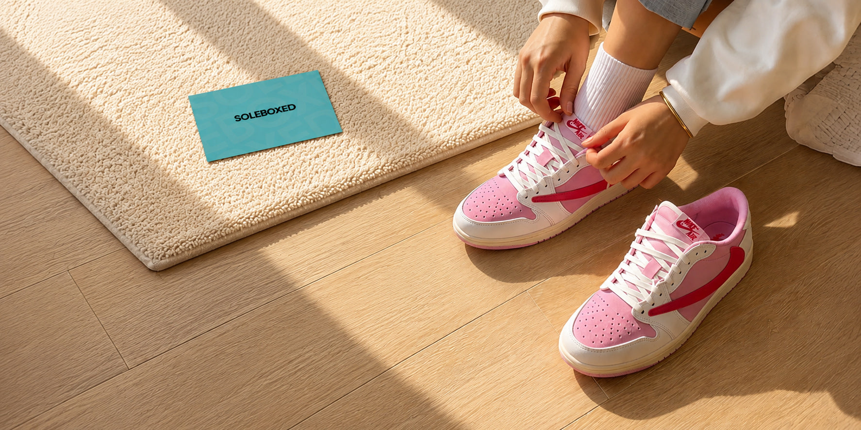 A person sitting on a wooden floor is tying the laces of a pair of pink and white low-top sneakers. A beige textured rug with a blue SOLEBOXED card lies nearby.