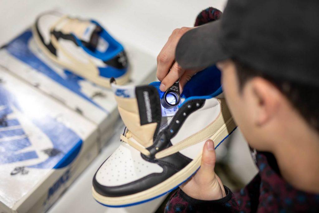 A close-up lifestyle shot of a person holding a high-top sneaker with a built-in camera lens on the tongue, inspecting the interior. The shoe is cream, black, and blue, and another shoe and its box are visible in the background.