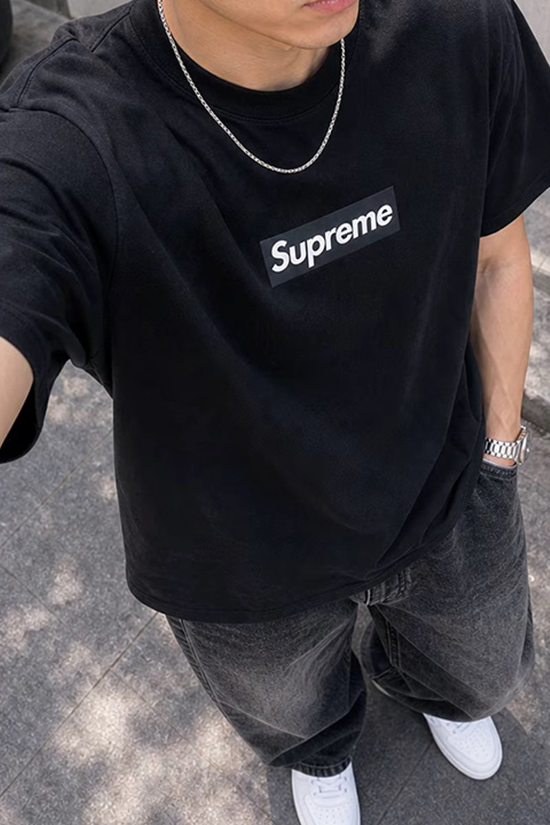 A person wearing a black Supreme box logo t-shirt, dark gray washed jeans, and white sneakers, photographed in a casual outdoor setting.