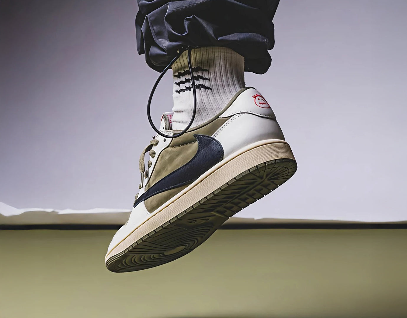 A single high-top sneaker worn with white socks and dark pants, suspended in mid-air against a soft gray background.
